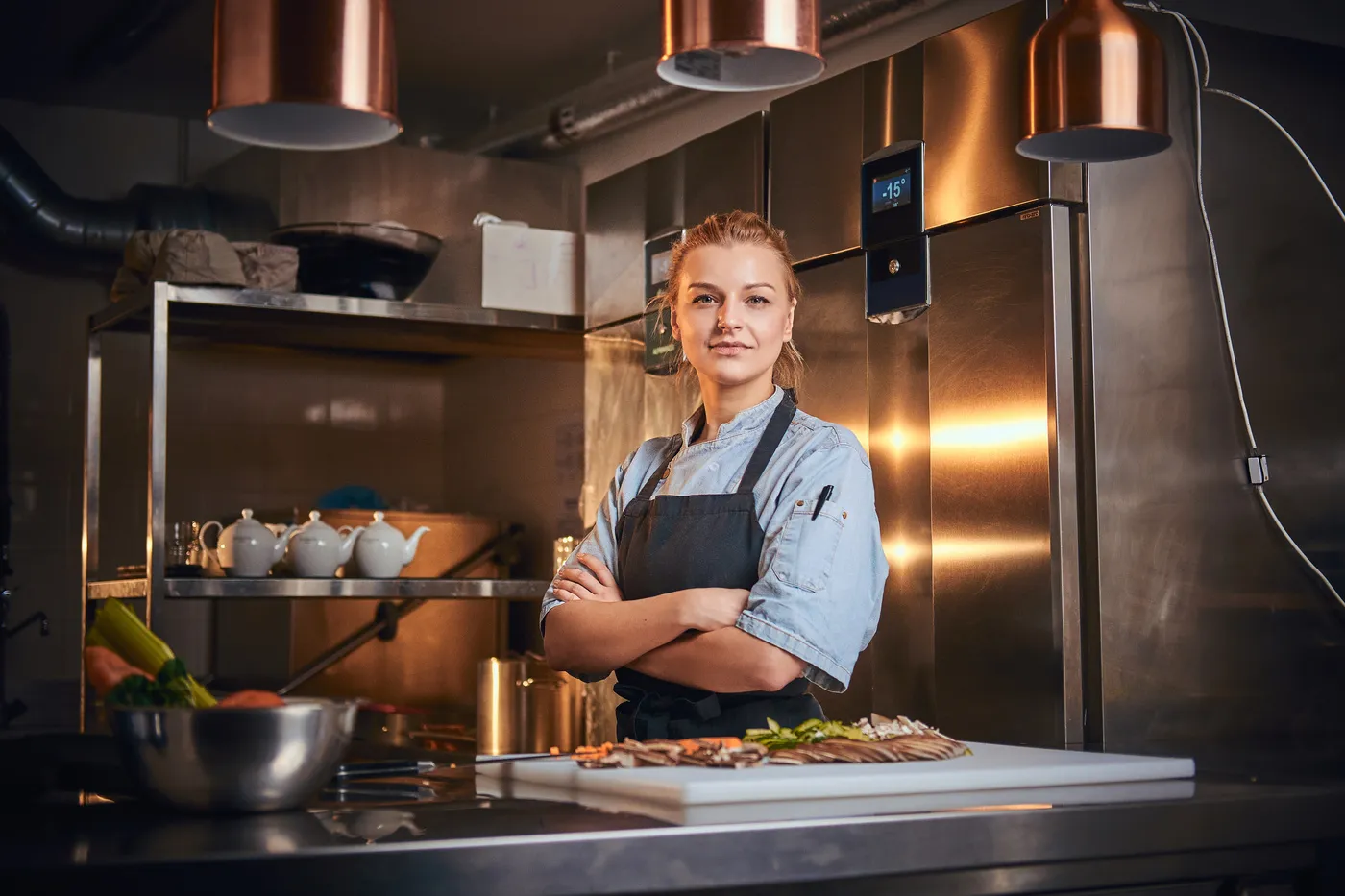Reinhardt koch - Kochkurse und Gastronomie-Service - Selbstbewusste und konzentrierte Köchin steht mit verschränkten Händen in einer dunklen Küche neben einem Schneidebrett mit Gemüse. Sie trägt eine Schürze und ein Jeanshemd und blickt in die Kamera – typischer Kochshow-Look.