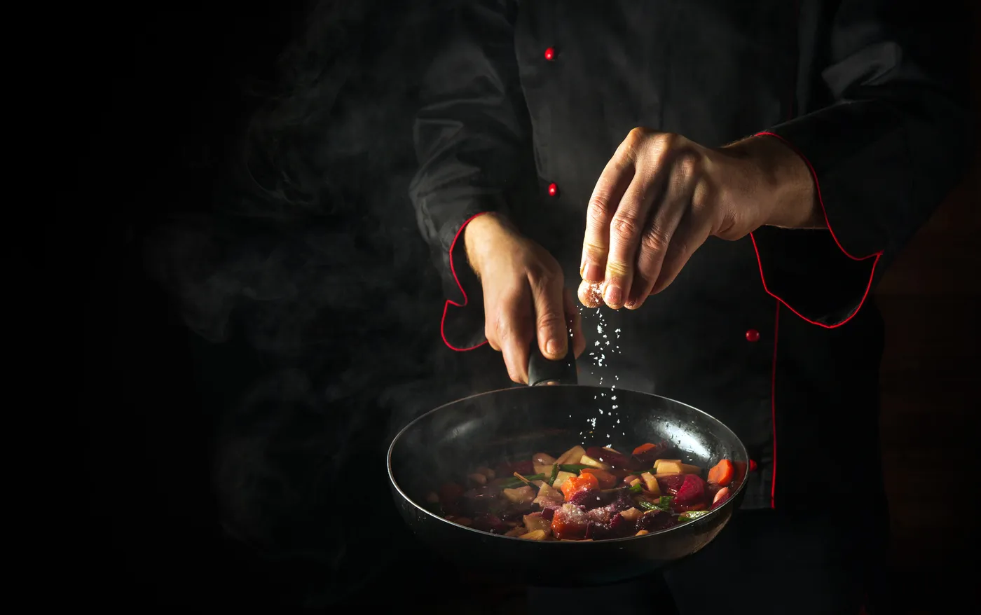 Reinhardt koch - Kochkurse und Gastronomie-Service - Zubereitung von frischem Gemüse. Der Koch gibt Salz in eine dampfend heiße Pfanne. Das Konzept der europäischen Küche für ein Hotel mit Werbefläche.
